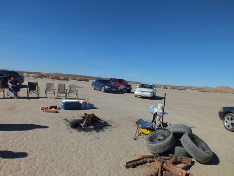 2003 Ford Crown Victoria - Fire pit and cooler ringed by folding chairs, stacked tires nearby and several cars across the lakebed.