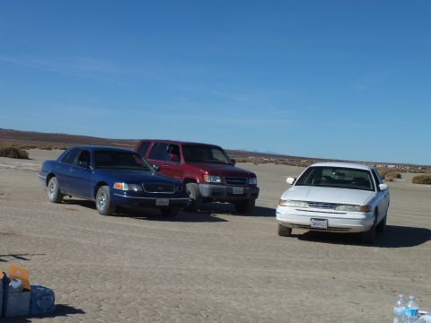 2003 Ford Crown Victoria - blue Crown Victoria, maroon SUV and white sedan line up on a cracked desert lakebed with brush behind.