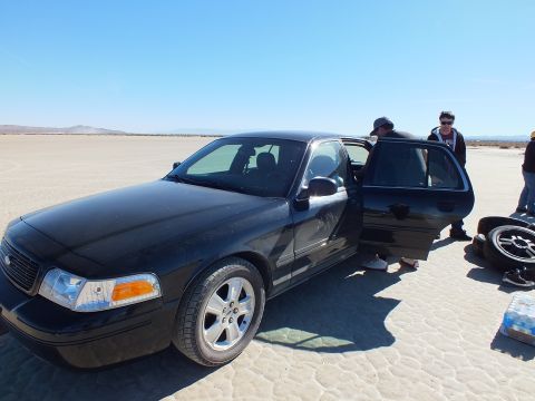 2003 Ford Crown Victoria - People stand by a black Crown Victoria with a rear door open, handling tires and water cases on the lakebed.
