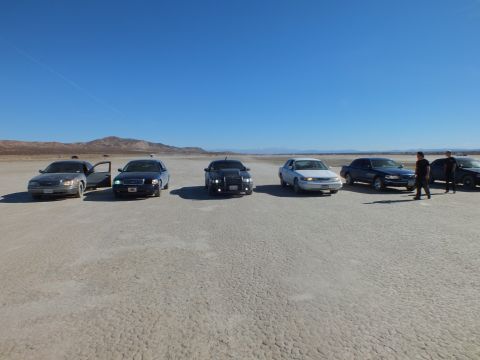 2003 Ford Crown Victoria - Line of parked sedans, among them Crown Victorias and a Dodge Charger, stretched across a wide desert lakebed.