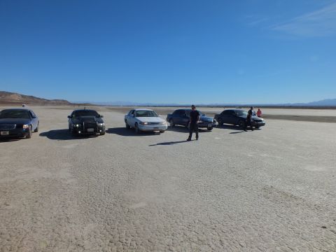 2003 Ford Crown Victoria - Multiple dark and light sedans line up across the lakebed as a person walks toward the camera.