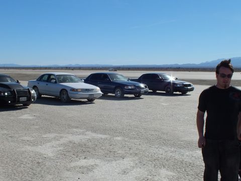 2003 Ford Crown Victoria - White and dark sedans plus a Dodge Charger line up on the lakebed; a man in a black shirt stands in the foreground.