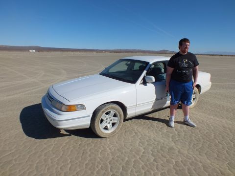 2003 Ford Crown Victoria - Man stands beside a white Crown Victoria parked on flat desert sand under clear blue sky