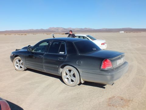 2003 Ford Crown Victoria - Rear quarter of a dusty black Crown Victoria with person beyond it on an open dry lakebed