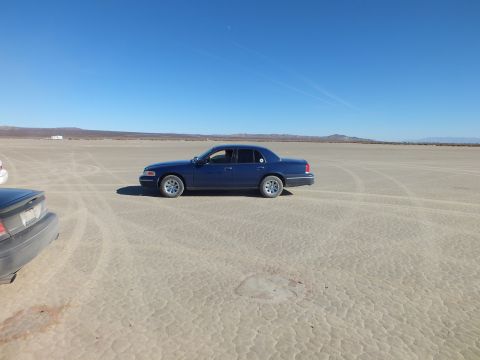 2003 Ford Crown Victoria - Side profile of a blue Crown Victoria on a dry lakebed, dark coupe partly visible at left