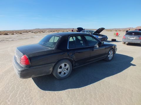 2003 Ford Crown Victoria - Rear quarter of a black Crown Victoria parked on desert sand, several vehicles with open hoods behind