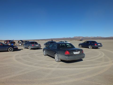 2003 Ford Crown Victoria - Multiple sedans gathered on a dry lakebed with sweeping tire tracks in the sand