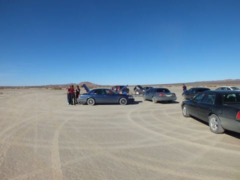 2003 Ford Crown Victoria - People stand around a blue sedan with its hood up on a dry lakebed, other cars parked nearby