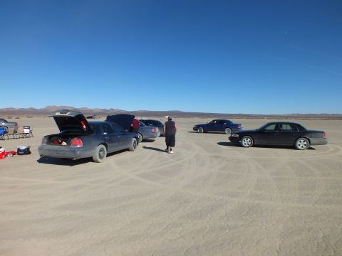 2003 Ford Crown Victoria - Group gathering on a dry lakebed with sedans open and camp chairs set out, person walking