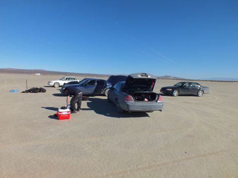 2003 Ford Crown Victoria - Man works at the open trunk of a gray sedan beside a red cooler on a flat dry lakebed