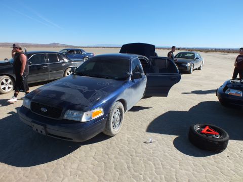 2003 Ford Crown Victoria - Blue Crown Victoria with door and trunk open beside a spare tire, people around it on a lakebed