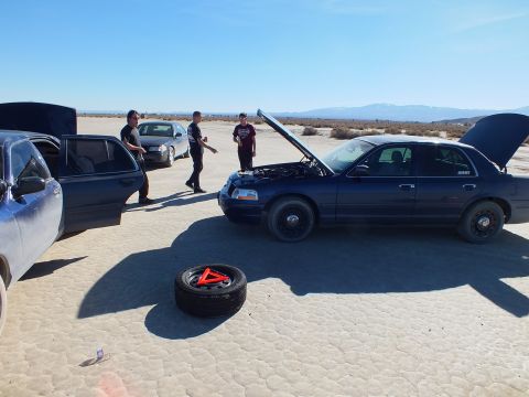 2003 Ford Crown Victoria - Men stand near a blue Crown Victoria with hood up and a spare tire on the ground, dry lakebed