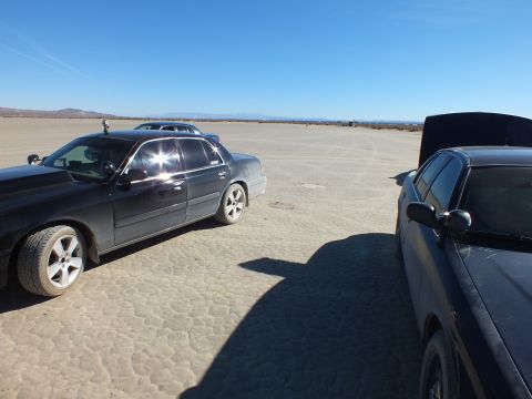 2003 Ford Crown Victoria - Black sedan with sun glare on windows parked beside a dark car on a desert dry lakebed