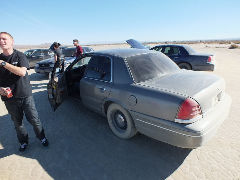 2003 Ford Crown Victoria - Man with a drink stands by a dusty gray Crown Victoria, doors open, on a flat dry lakebed