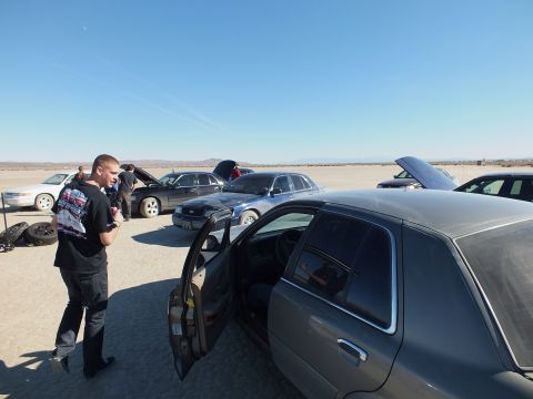 2003 Ford Crown Victoria - Young man stands by a gray Crown Victoria with door open, more cars behind on the dry lakebed