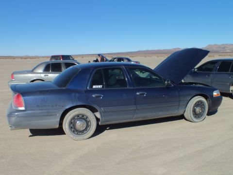 2003 Ford Crown Victoria - Hood raised on a dusty blue Crown Victoria, with more sedans and a pickup lined up across the playa.