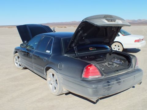 2003 Ford Crown Victoria - Black Crown Victoria opened front and rear beside a white sedan on the cracked desert surface.