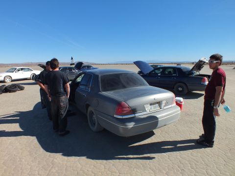 2003 Ford Crown Victoria - Three men stand by a dusty silver Crown Victoria as several sedans sit with hoods raised behind them.