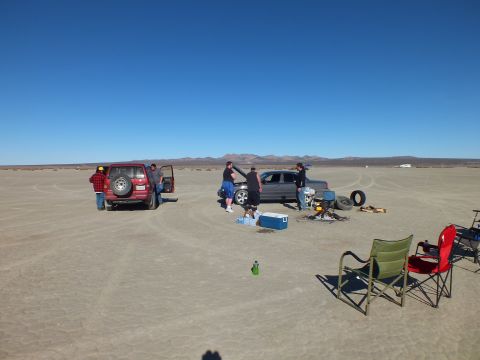 2003 Ford Crown Victoria - People gather by two SUVs with coolers, chairs and a tire on an open desert flat ringed by mountains.