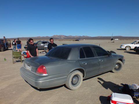 2003 Ford Crown Victoria - Dusty gray Crown Victoria sits among chairs and other sedans on a mountain-ringed lakebed.