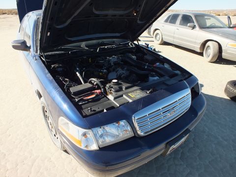 2003 Ford Crown Victoria - Open hood reveals the V8 of a blue Crown Victoria, a silver sibling parked behind on the lakebed.