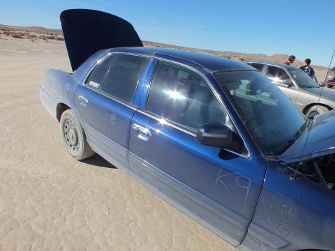 2003 Ford Crown Victoria - Dust-scrawled POLICE and K9 markings cover a blue Crown Victoria with its hood raised on the playa.