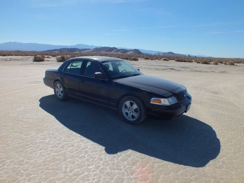 2003 Ford Crown Victoria - Black Crown Victoria casts a long shadow on the lakebed with a mountain range on the horizon.