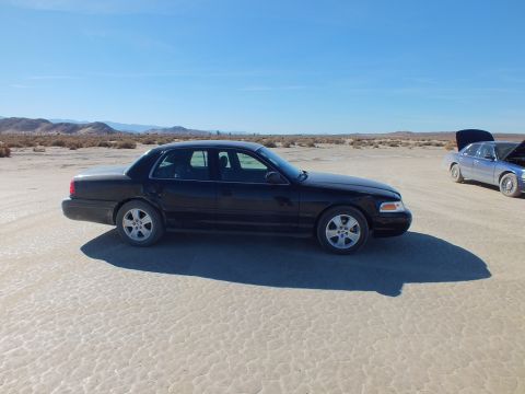 2003 Ford Crown Victoria - Profile of a black Crown Victoria on the playa; a silver sedan sits with its hood up in the distance.