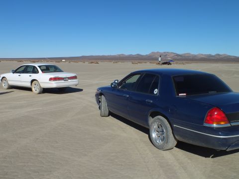 2003 Ford Crown Victoria - dark blue Crown Victoria and a white sibling sit on the cracked playa beneath distant ranges.