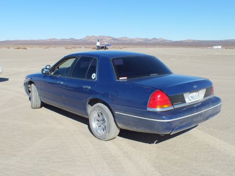 2003 Ford Crown Victoria - Dark blue Crown Victoria with chrome wheels on the playa, Ford oval and taillights at the rear.