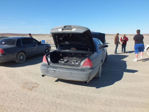 2003 Ford Crown Victoria - Trunk-up rear view of a charcoal sedan on cracked desert playa, a second dark car at left.