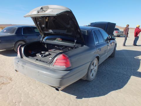 2003 Ford Crown Victoria - Rear three-quarter of a dark full-size sedan, trunk lid raised, other cars and a hill behind.