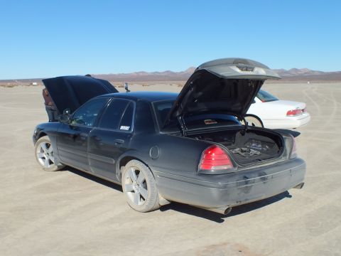 2003 Ford Crown Victoria - Rear quarter of a black full-size sedan, hood and trunk up, white car parked to the right.