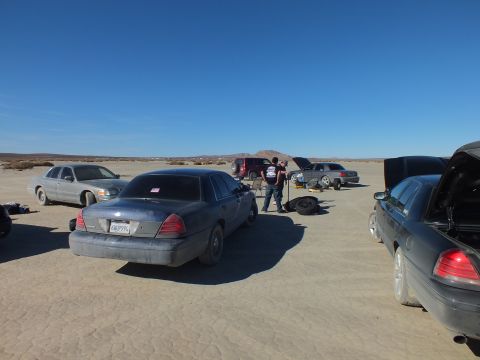 2003 Ford Crown Victoria - Rear of a dusty gray sedan with people and tires beyond, several cars on the playa.