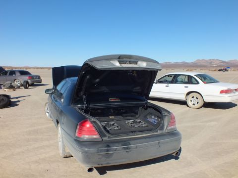 2003 Ford Crown Victoria - Trunk-up dark sedan with subwoofer box, white full-size sedan parked behind on the playa.