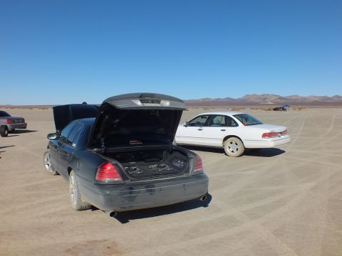2003 Ford Crown Victoria - Rear of a dark sedan, trunk open, a white sedan and distant mountains across the playa.