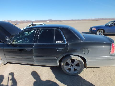 2003 Ford Crown Victoria - Side view of a black sedan, hood raised, another sedan parked in the distance on playa.