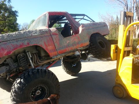 1978 Ford Bronco - A mud-covered red 1978 Ford Bronco is lifted by a yellow forklift, its off-road modifications.