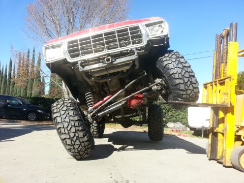 1978 Ford Bronco - Front end of a red-and-white lifted Ford 4x4 hoisted in the air on a yellow forklift, exposing the Dana front axle with red-painted differe…