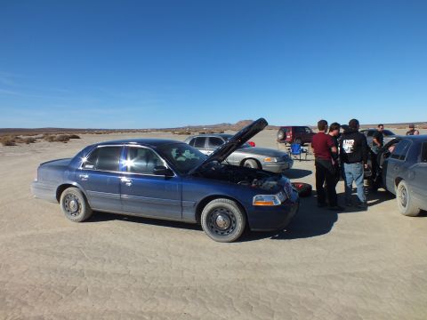 2003 Ford Crown Victoria - Front quarter of a blue sedan, hood up, people gathered, more cars on the cracked playa.