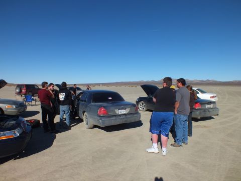 2003 Ford Crown Victoria - People cluster between several full-size sedans on cracked playa under a clear blue sky.