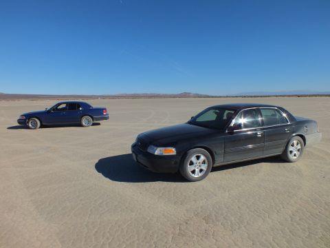 2003 Ford Crown Victoria - Black sedan in foreground and a dark blue sedan beyond, spread across cracked playa.