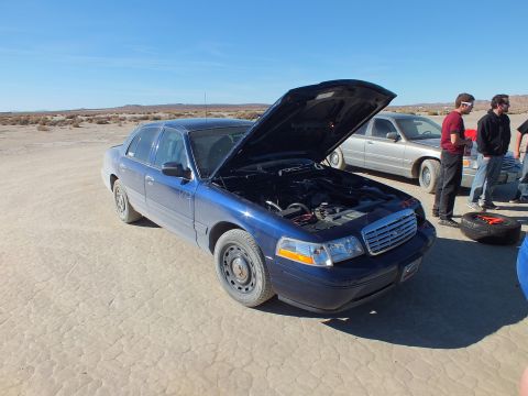 2003 Ford Crown Victoria - Front three-quarter of a blue Ford sedan, hood raised, spare tire and people at right.