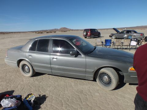 2003 Ford Crown Victoria - Side profile of a dusty gray sedan, camp chairs and an SUV across the cracked playa.