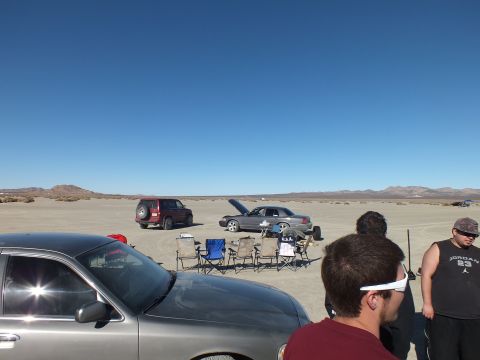 2003 Ford Crown Victoria - Group of people and parked cars with camp chairs spread across the cracked desert flats.