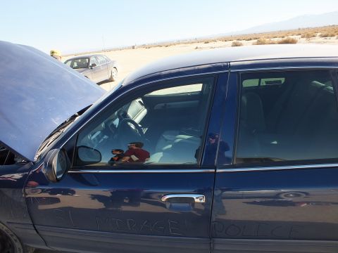 2003 Ford Crown Victoria - Side window and door of a blue sedan, hood up, EL MIRAGE POLICE scrawled in the dust.