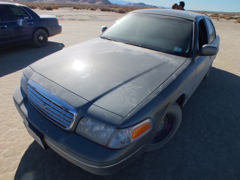 2003 Ford Crown Victoria - Dusty gray Ford sedan with Ford oval grille, blue car behind on the cracked playa.