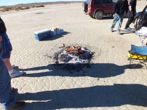 2003 Ford Crown Victoria - Spent fire ring with ash on cracked playa, coolers and an SUV behind, people standing by.
