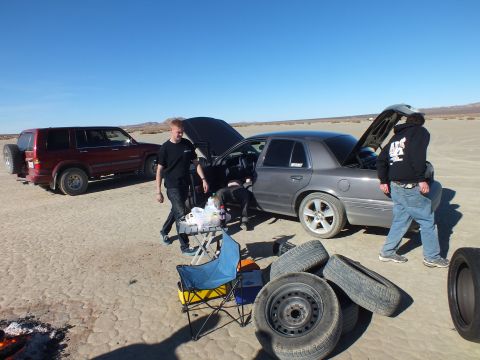 2003 Ford Crown Victoria - Gray full-size sedan with hood and trunk up, steel wheels stacked beside it on the playa.