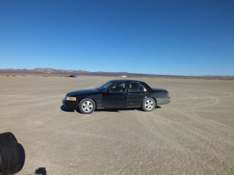 2003 Ford Crown Victoria - Side profile of a black sedan alone on cracked playa, tire tracks and mountains beyond.